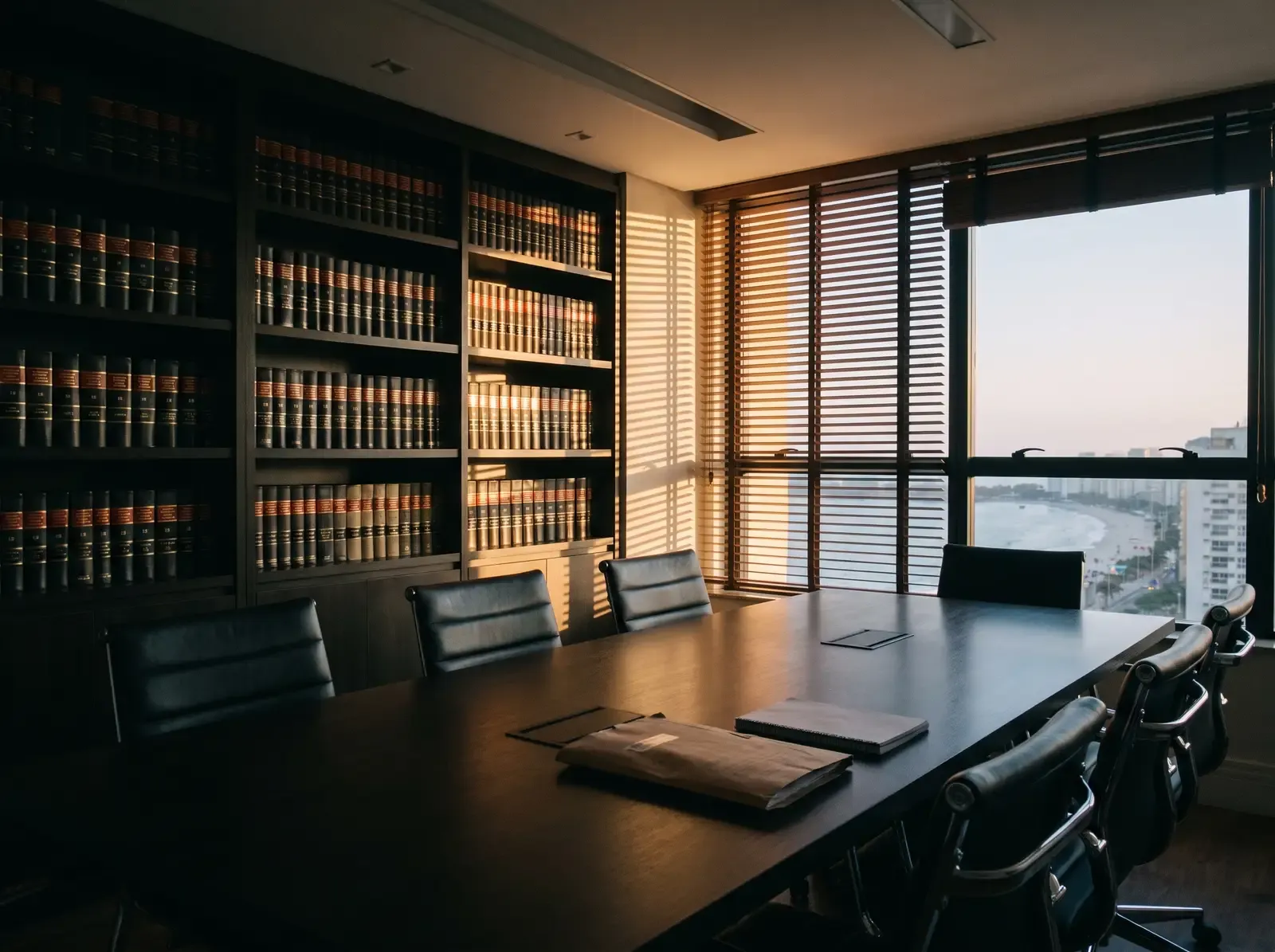 Sala de reunião de escritório de advocacia com mesa de madeira escura, cadeiras de couro e estante de livros jurídicos, iluminada pela luz da janela